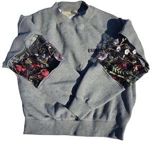 May include: A grey crewneck sweatshirt featuring black mesh panels on the upper sleeves adorned with colourful floral embroidery. The chest displays the black text ESSENTIALS FEAR OF GOD. The garment has ribbed cuffs and a ribbed hemline.