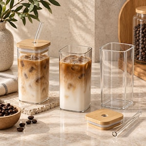 May include: Three square-shaped clear glass tumblers. Two tumblers are filled with iced coffee and milk, each topped with a square bamboo lid and a clear glass straw. One empty tumbler sits beside a separate bamboo lid and a clear glass straw.