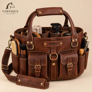 May include: A brown leather grooming bag featuring two front pockets with buckle closures, a central ring, and a nameplate engraved with the name Willow. The bag includes a top handle, a detachable shoulder strap, and multiple interior compartments.
