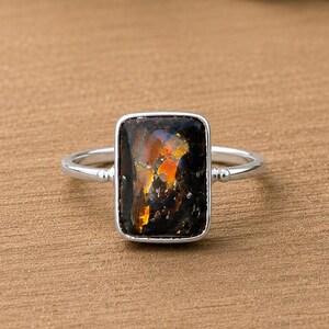Op de afbeelding: Een zilveren ring met een rechthoekige, donkergekleurde edelsteen met iriserende oranje en gele insluitingen. De steen is gezet in een gladde metalen zetting en de dunne, gepolijste ringband is verbonden met de zetting via kleine, ronde accenten.