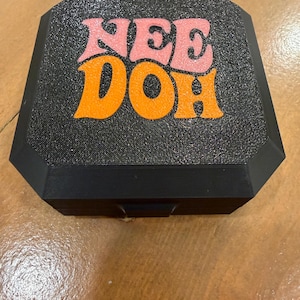 May include: A black, octagonal storage box with a textured, glittery finish. The top surface features the text "NEE DOH" in bold, stylized lettering, with "NEE" in light pink and "DOH" in bright orange, centered on the lid.