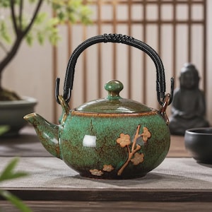Pode incluir: Uma chaleira de cerâmica redonda com esmalte verde manchado e acabamento castanho escuro perto da base. A chaleira apresenta um relevo de um ramo florido com flores castanho-claras e uma pega preta enrolada presa a dois anéis metálicos.