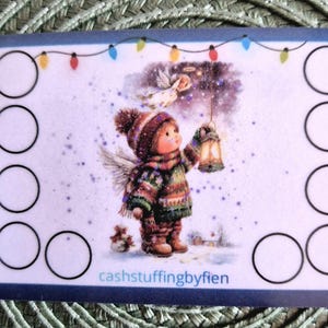 Op de afbeelding: Een rechthoekige witte kaart met een centrale illustratie van een kind in winterkleding dat een lantaarn vasthoudt, met een kleine engel erboven. De kaart heeft een snoer met gekleurde kerstverlichting bovenaan, tien zwarte cirkels en de tekst cashstuffingbyfien.