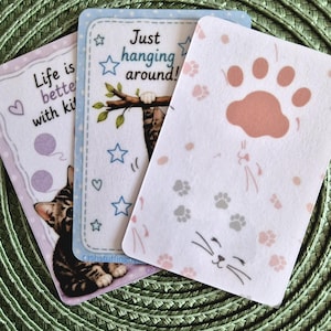 Op de afbeelding: Drie rechthoekige kaarten met illustraties met kattenthema. Eén kaart toont een kitten met de tekst Life is better with kittens en garenbollen. Een andere toont een kat die aan een tak hangt met de tekst Just hanging around. De derde heeft pootafdrukken.