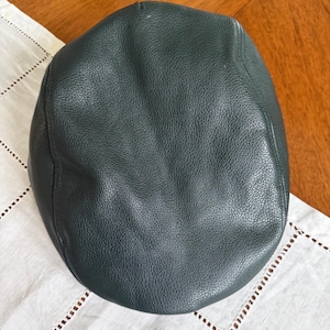 May include: A dark forest green flat cap crafted from textured leather. The cap features a rounded, structured crown with visible stitching along the side panels and a smooth, pebbled surface texture throughout the entire exterior of the headwear.