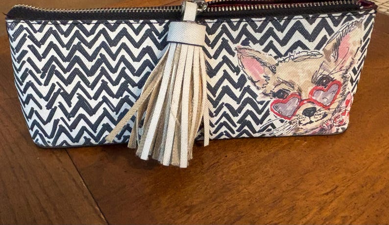 Puede incluir: Dos neceseres con cremallera. Una bolsa presenta un patr&oacute;n de chevr&oacute;n en blanco y negro, la otra tiene un chihuahua con gafas de sol en forma de coraz&oacute;n. Ambas bolsas tienen una borla beige adjunta a la cremallera.