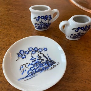 May include: A white porcelain miniature tea set with blue floral and pagoda designs. The set includes two small pitchers and a saucer. The blue design is hand-painted on the white porcelain. The set is displayed on a wooden surface.