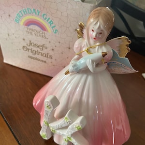 May include: A Josef Originals figurine of a girl in a pink and white dress, holding a blue object. The figurine has gold wings and a tag that says "Birthday Girls". The box behind the figurine says "Birthday Girls Through the Years".