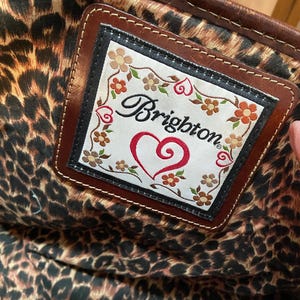 Puede incluir: Primer plano de una etiqueta de cuero marr&oacute;n con el logotipo de Brighton, un coraz&oacute;n rojo y detalles florales. La etiqueta se encuentra sobre un tejido interior con estampado de leopardo, posiblemente de un bolso.