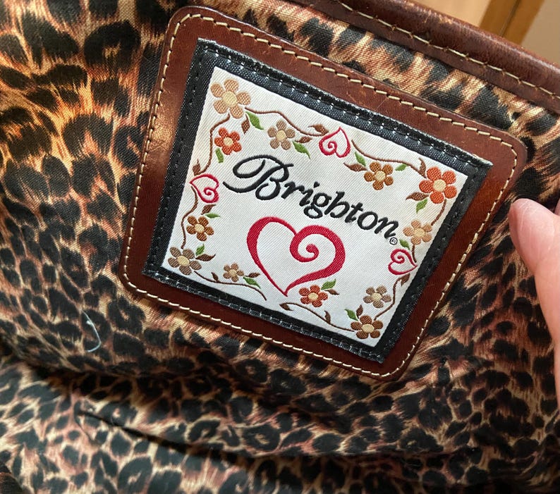 Puede incluir: Primer plano de una etiqueta de cuero marr&oacute;n con el logotipo de Brighton, un coraz&oacute;n rojo y detalles florales. La etiqueta se encuentra sobre un tejido interior con estampado de leopardo, posiblemente de un bolso.