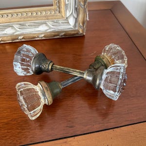 May include: Two clear, fluted glass door knobs with metallic bases and threaded spindles. The knobs feature a faceted, multi-sided design and are connected by a central metal rod. The hardware shows signs of age with a tarnished, dark finish.