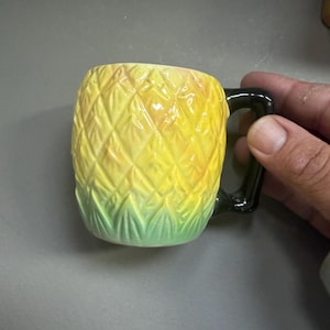 May include: A pineapple-shaped ceramic mug. The mug has a textured yellow body with green and yellow leaves at the base and a black handle. The design of the mug is intended to look like a pineapple.