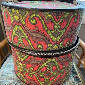May include: Vintage two-piece hatbox set. The hatboxes are cylindrical with a black handle and trim. The fabric is patterned with red, green, and yellow paisley designs. The bottom hatbox has a zipper closure. The set is sitting on a chair.