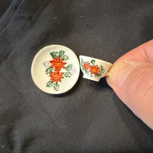 May include: A miniature white ceramic tea set consisting of a small saucer and a matching teacup. Both pieces feature a hand-painted floral pattern with vibrant orange-red flowers and green leaves. The teacup has a thin gold-colored rim.