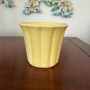 Peut inclure: Un cache-pot en céramique jaune avec un bord évasé et des détails cannelés verticaux le long du corps extérieur. Le pot présente une finition lisse et brillante ainsi qu'une base circulaire, offrant un design simple pour contenir des plantes.