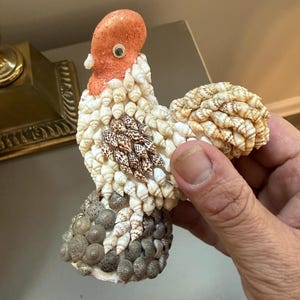 May include: A decorative cockerel figurine crafted from various small seashells. The head is a textured orange material with a single googly eye. The body is covered in white, brown, and grey shells, with a base composed of small, round grey shells.