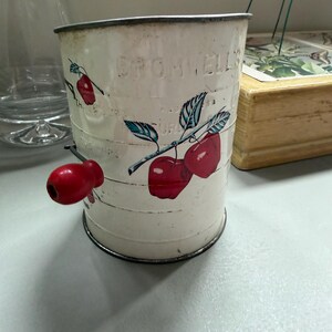 以下が含まれることがあります： 赤い木製のハンドルが付いた、ヴィンテージの白い金属製のキッチン用品。赤いサクランボと緑の葉で飾られています。「BROMWELL'S」の文字が側面にエンボス加工されています。キッチン装飾用の魅力的な作品です。