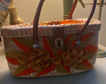 Könnte beinhalten: Eine geflochtene Strohtasche mit einem braunen Ledergriff und einem Verschluss. Die Tasche ist mit orangefarbenen und braunen Stoffblumen verziert.