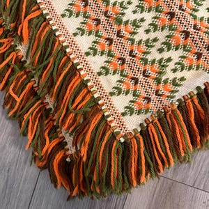 May include: A woven textile throw featuring a cream-colored base with a repeating geometric floral pattern in shades of orange, dark brown, and olive green. The edges are finished with long, thick fringe in alternating orange, brown, and green yarn.