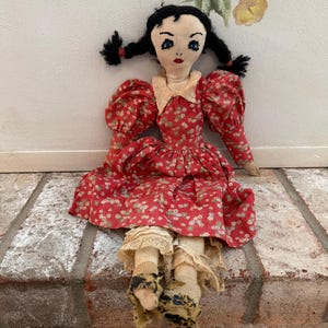 May include: A cloth doll with black yarn hair styled in pigtails, featuring embroidered blue eyes and red lips. The doll wears a red dress with a small white floral pattern, a cream-colored collar, and layered lace-trimmed undergarments with dark patterned footwear.