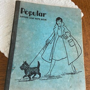 May include: A blue notebook with the text "Popular Loose Leaf Note Book" on the cover. The cover features a black and white illustration of a woman in a coat walking a dog.