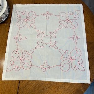 May include: White fabric square with a red embroidered design of repeating floral patterns.