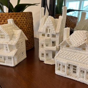 May include: Three white plastic canvas houses of varying sizes sit on a dark wooden surface. The houses have detailed architectural features, including windows, doors, and chimneys. The houses are decorative and appear to be handmade.