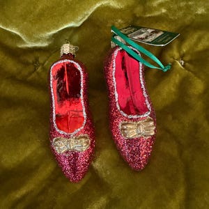 May include: Two red glass shoe ornaments covered in fine glitter, each featuring a metallic gold bow at the toe and a silver glitter trim around the opening. One ornament includes a green ribbon loop for hanging and a small paper product tag.