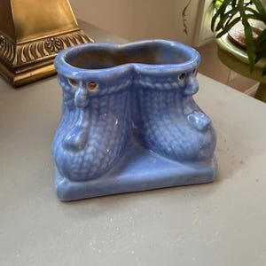Puede incluir: Una maceta de cerámica azul claro con forma de un par de botas unidas con un patrón de tejido texturizado. La maceta presenta una base lisa, una abertura ancha en la parte superior y pequeños orificios circulares cerca del tobillo.