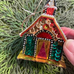 Puede incluir: Adorno navideño hecho a mano con forma de iglesia. El adorno está adornado con lentejuelas doradas, rojas, verdes y bronce. El tejado es rojo con un campanario blanco y negro. La puerta es dorada con un triángulo rojo.