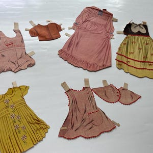 May include: A selection of vintage paper doll outfits, including dresses, a jacket, and accessories. The dresses are in various colours, such as pink, yellow, and red, with decorative details. The paper dolls have tabs for attachment.