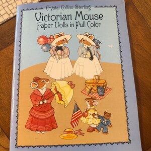 May include: A softcover book titled Victorian Mouse Paper Dolls in Full Color by Crystal Collins-Sterling. The light blue cover features illustrations of anthropomorphic mice dressed in period clothing, with accessories like balloons, a picnic basket, a yellow parasol, and an American flag.