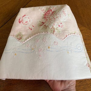 Peut inclure: Un chauffe-thé fait à la main avec un motif floral de roses roses et de feuilles vertes sur un fond rose clair. La partie inférieure est blanche avec des détails floraux brodés et un bord festonné.