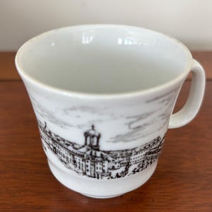 Puede incluir: Taza de té de cerámica blanca con una ilustración arquitectónica en blanco y negro. La taza tiene un asa pequeña y un borde ligeramente acampanado. El diseño representa un paisaje urbano detallado.