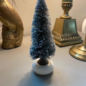 Può includere: Un piccolo albero di Natale artificiale floccato con una base bianca. L'albero è decorato con glitter bianco.