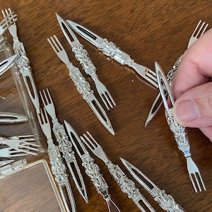 Puede incluir: Colección de tenedores de aperitivo plateados y ornamentados con mangos decorativos. Algunos tenedores están colocados sobre una superficie de madera, otros en un recipiente de plástico transparente. Diseño clásico y elegante.