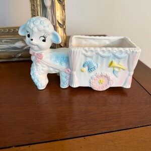 May include: A ceramic planter shaped like a lamb pulling a cart. The lamb is white with blue accents and a pink bow. The cart is white with blue scalloped trim, featuring a blue striped hat, a yellow trumpet, and a pink flower with a yellow centre.