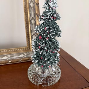 Op de afbeelding: Een kunstmatige miniatuurkerstboom met donkergroene naalden, witte vlokken die sneeuw nabootsen en kleine rode bessen. De boom is bevestigd op een ronde, heldere voet van geperst glas met een gestructureerde, geschulpte rand.