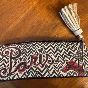 Puede incluir: Un estuche rectangular con un patr&oacute;n de zigzag en blanco y negro. La palabra "Par&iacute;s" est&aacute; escrita en rojo. La Torre Eiffel se muestra en rojo. Una borla beige cuelga de la cremallera.