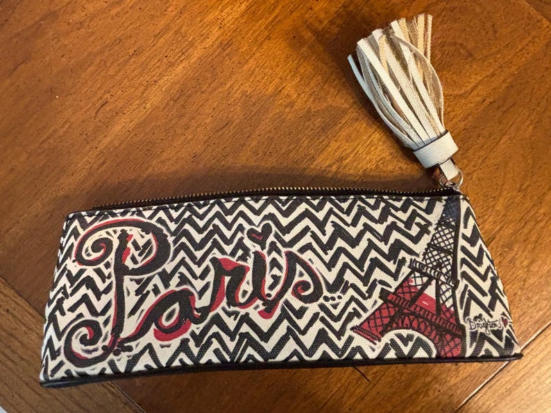 Puede incluir: Un estuche rectangular con un patr&oacute;n de zigzag en blanco y negro. La palabra "Par&iacute;s" est&aacute; escrita en rojo. La Torre Eiffel se muestra en rojo. Una borla beige cuelga de la cremallera.