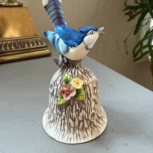 以下が含まれることがあります： 上にアオカケスがとまっている陶器製のベル。ベルは茶色の樹皮のディテールが施された質感のある切り株の形をしており、前面にはピンクと黄色の花と緑の葉の小さな房が装飾されています。