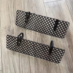 May include: Two rectangular black metal shelves featuring a repeating quatrefoil cutout pattern. Each shelf has two integrated black metal hooks on the back for mounting purposes, providing a structured and open design for storage or display.