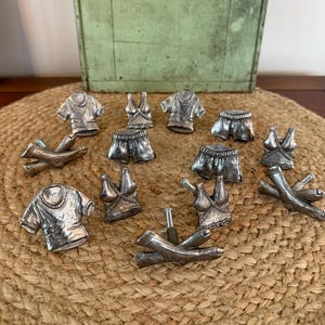 May include: A collection of silver-toned metal cabinet knobs shaped like various articles of clothing, including t-shirts, tank tops, shorts, and pants. Each piece features a metallic finish with detailed textures representing fabric folds and seams.