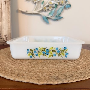 May include: A square white milk glass baking dish featuring a decorative pattern of blue berries and green leaves along the exterior side. The dish has a smooth, glossy finish with a wide rim and a shallow, open interior design for food preparation.