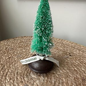 May include: A small green bottle brush Christmas tree with white flocking on a brown base. The tree is decorated with a white ribbon that says "Merry Christmas".
