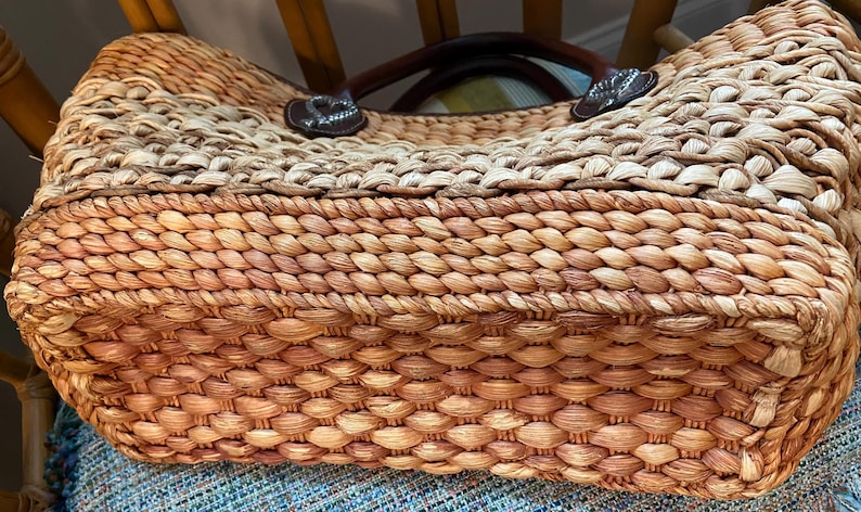 Puede incluir: Bolso tote de paja tejida con asas de cuero marr&oacute;n y detalles decorativos. El bolso presenta una paleta de colores naturales con un dise&ntilde;o tejido apretado, mostrando una estructura robusta y espaciosa. El bolso est&aacute; dise&ntilde;ado para transportar objetos.