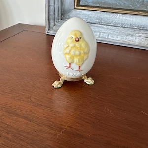 Puede incluir: Un huevo de cerámica blanca con un diseño de pollito amarillo y el año 1972 impreso en la parte inferior. El huevo está sobre un soporte de metal dorado con tres patas.