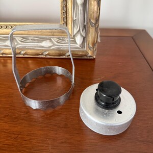 May include: A metal biscuit cutter with a scalloped edge and a tall handle, positioned next to a circular metal lid featuring a small black knob and a single ventilation hole. Both items are constructed from silver-toned metal components.