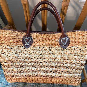 Puede incluir: Bolso tote de paja tejida con asas y ribetes de cuero marr&oacute;n. El bolso presenta un patr&oacute;n de ondas repetidas y detalles decorativos en forma de coraz&oacute;n en las fijaciones de las asas. El bolso tiene forma rectangular.