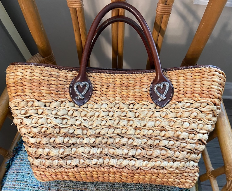 Puede incluir: Bolso tote de paja tejida con asas y ribetes de cuero marr&oacute;n. El bolso presenta un patr&oacute;n de ondas repetidas y detalles decorativos en forma de coraz&oacute;n en las fijaciones de las asas. El bolso tiene forma rectangular.